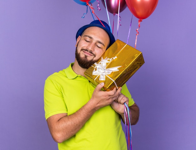 Heureux avec les yeux fermés, jeune homme portant un chapeau de fête tenant des ballons avec une boîte-cadeau isolée sur un mur bleu