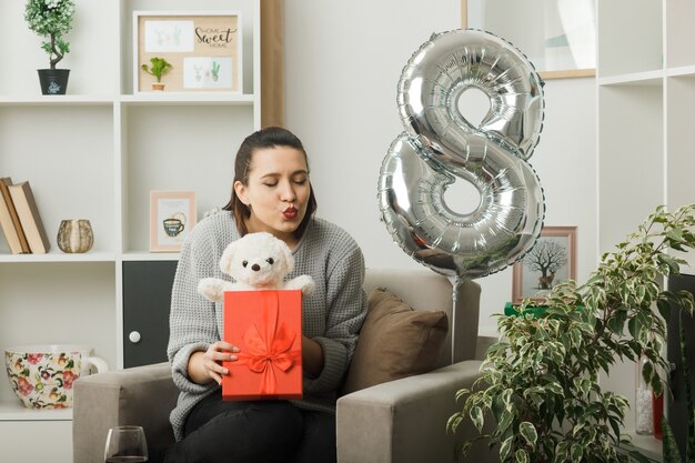 Heureux avec les yeux fermés montrant le geste du baiser belle fille le jour de la femme heureuse tenant un cadeau avec un ours en peluche assis sur un fauteuil dans le salon