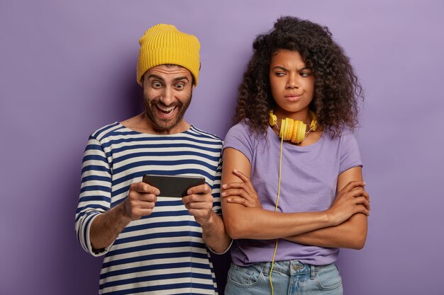 Un hipster accro joue au smartphone, ignore sa petite amie, la femme afro s'ennuie, garde les bras croisés et regarde négativement.