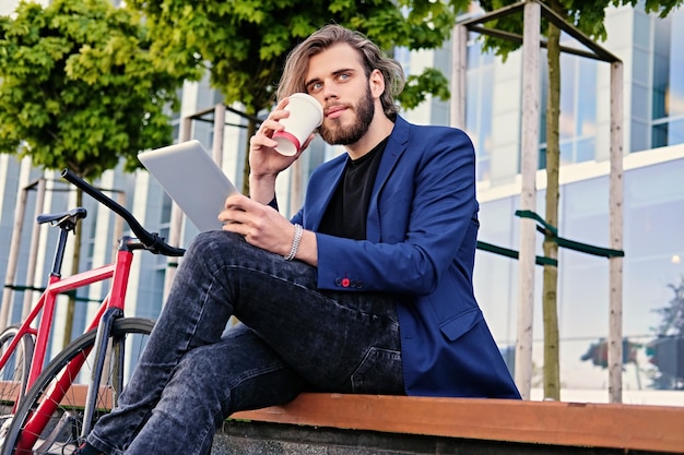 Un hipster barbu aux longs cheveux blonds vêtu d'un jean boit du café et tient une tablette PC avec un vélo fixe dans un parc en arrière-plan.