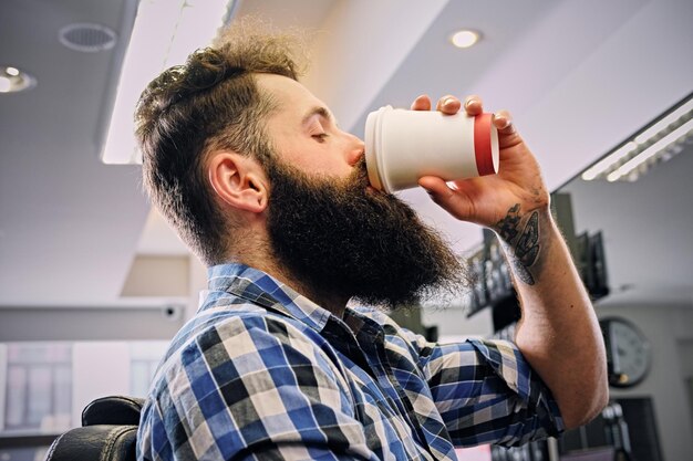 Un hipster barbu élégant vêtu d'une chemise polaire boit du café dans un salon de coiffure.