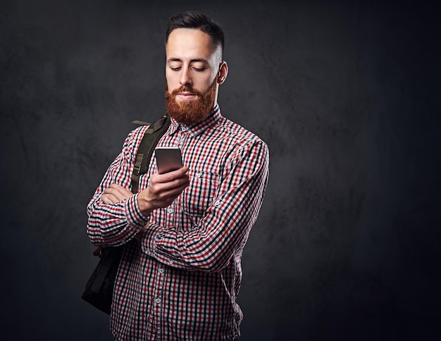 Hipster barbu rousse mâle dans une chemise polaire rouge détient smartphone sur fond gris.