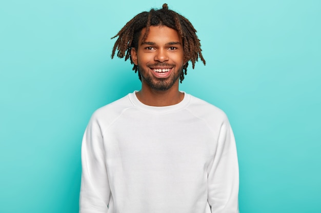 Photo gratuite hipster avec des dreadlocks, sourit largement, montre des dents, porte un pull blanc décontracté, satisfait après un week-end avec des amis