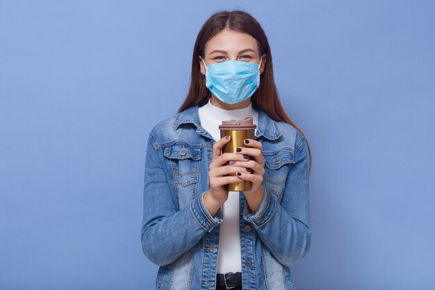 Hipster girl in medical face mask and denim jacket drink coffee from thermo mug