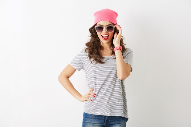 Hipster jolie femme au chapeau rose, lunettes de soleil, souriant, isolé, dents blanches, lèvres rouges, cheveux bouclés, portant un t-shirt