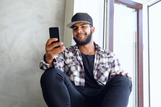 Un hipster noir vêtu d'une chemise polaire et d'une balle de baseball est assis près de la fenêtre et utilise un smartphone.