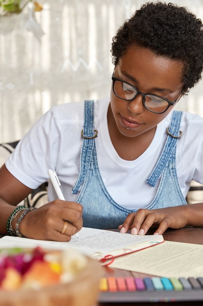 Hipster à la peau sombre dans les lunettes, écrit dans un cahier, fait ses devoirs