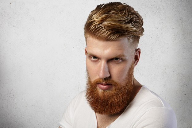 Hipster rousse avec coupe de cheveux à la mode et barbe floue vêtu d'un t-shirt blanc posant à l'intérieur, ayant un look sérieux et maussade.