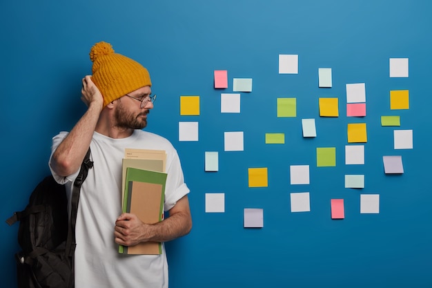 Un hipster sérieux se frotte la tête, regarde sérieusement les notes colorées collées au mur, rappelle les informations nécessaires