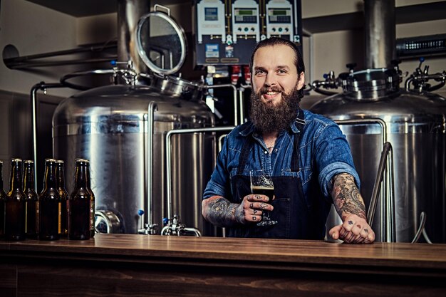 Un hipster tatoué barbu souriant dans une chemise en jean et un tablier travaillant dans une usine de brasserie, debout derrière un comptoir, tient un verre de bière pour le contrôle de la qualité.