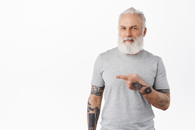 Hipster vieil homme avec une longue barbe montrant une publicité et souriant heureux, recommandant une entreprise, debout sur un mur blanc