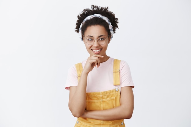 Hmm super concept, faisons-le. Heureux séduisant jeune blogueuse internet afro-américaine femme dans des verres, bandeau et salopette jaune, souriant et tenant la main sur le menton, acceptant le plan