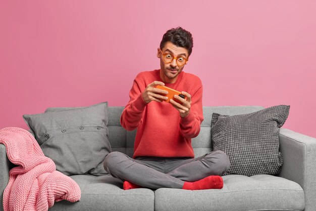 Un homme accro joue à des jeux en ligne sur smartphone est assis les jambes croisées sur un canapé confortable