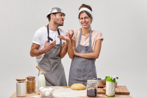 Un homme adulte agacé regarde sa femme avec colère, demande d'arrêter de cuisiner, se sent fatigué de faire de la pâte, une femme joyeuse en tablier aime le passe-temps et faire de délicieuses pâtisseries. Culinaire et pâtisserie pendant la quarantaine