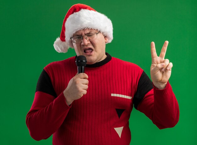 Homme adulte confiant portant des lunettes et bonnet de Noel parlant dans le microphone regardant la caméra faisant signe de paix isolé sur fond vert