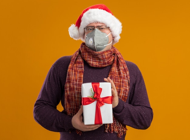 Homme adulte confiant portant des lunettes de masque de protection et bonnet de Noel avec foulard autour du cou tenant un paquet cadeau regardant la caméra isolée sur fond orange