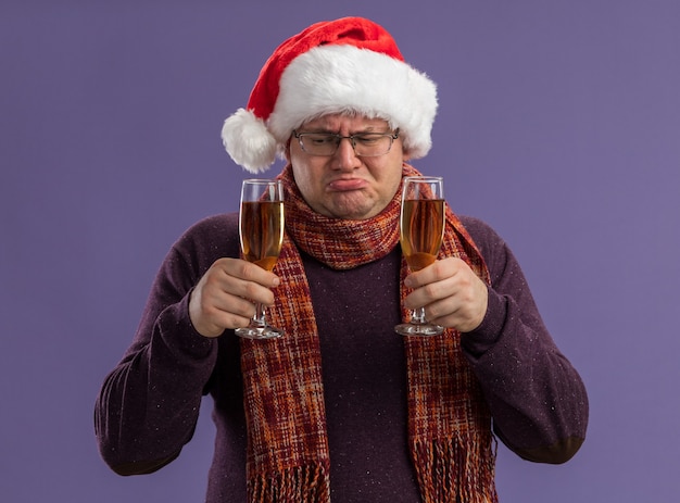 Homme adulte confus portant des lunettes et bonnet de Noel avec foulard autour du cou tenant deux verres de champagne en regardant un isolé sur fond violet