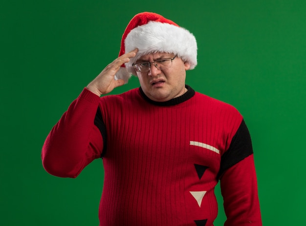 Homme adulte confus portant des lunettes et bonnet de Noel regardant la caméra toucher la tête isolé sur fond vert