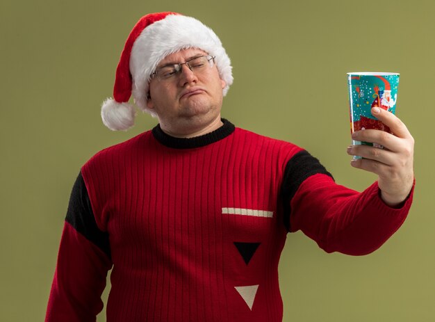 Homme adulte confus portant des lunettes et un bonnet de noel tenant une tasse de café de noël l'étirant et le regardant isolé sur un mur vert olive