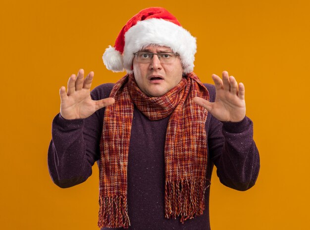 Homme adulte effrayé portant des lunettes et bonnet de Noel avec écharpe autour du cou regardant la caméra en gardant les mains en l'air isolé sur fond orange