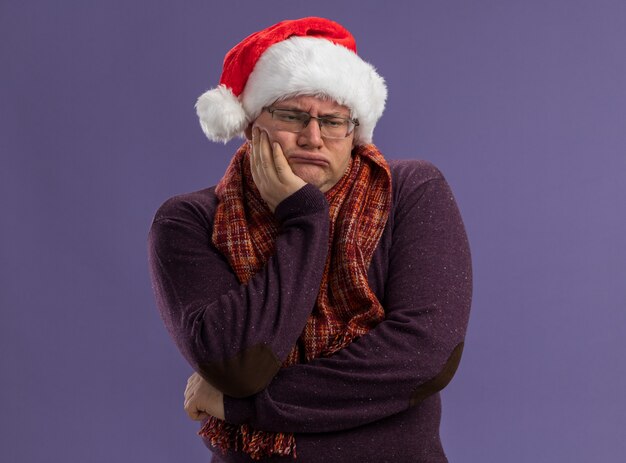 Homme adulte ennuyé portant des lunettes et un bonnet de noel avec une écharpe autour du cou en gardant la main sur le menton en regardant le côté isolé sur un mur violet