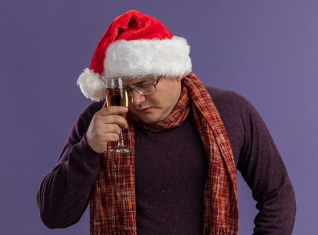 Homme adulte fatigué portant des lunettes et bonnet de Noel avec foulard autour du cou touchant le front avec verre de champagne avec les yeux fermés isolé sur fond violet