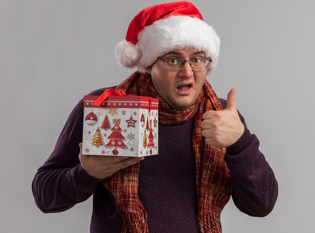 Homme adulte impressionné portant des lunettes et un bonnet de noel avec une écharpe autour du cou tenant un paquet cadeau de noël montrant le pouce vers le haut isolé sur un mur blanc