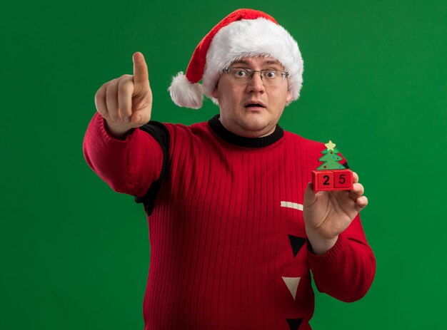 Homme adulte impressionné portant des lunettes et bonnet de noel tenant jouet d'arbre de Noël avec date à la recherche et pointant vers la caméra isolée sur fond vert