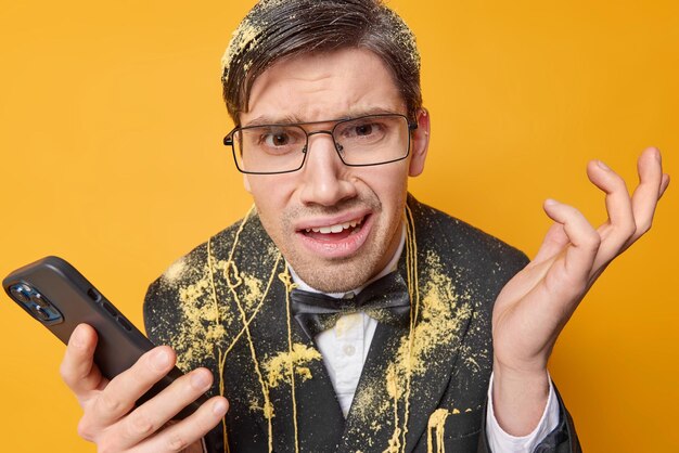 Un homme adulte indigné et indigné hausse les épaules se sent dérangé utilise un téléphone portable envoie des messages texte vêtu d'un costume formel festif porte des lunettes transparentes isolées sur fond jaune