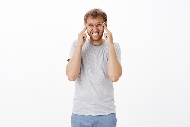 Homme adulte intense perplexe avec des poils dans des lunettes serrant les dents en plissant les yeux et en étirant les paupières pour voir clairement avoir des problèmes de vue tout en essayant de nouvelles lunettes dans un magasin d'opticien sur un mur blanc