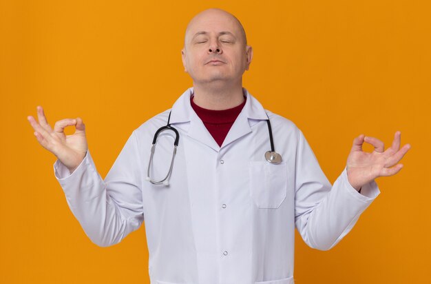 Homme adulte paisible en uniforme de médecin avec stéthoscope méditant debout les yeux fermés