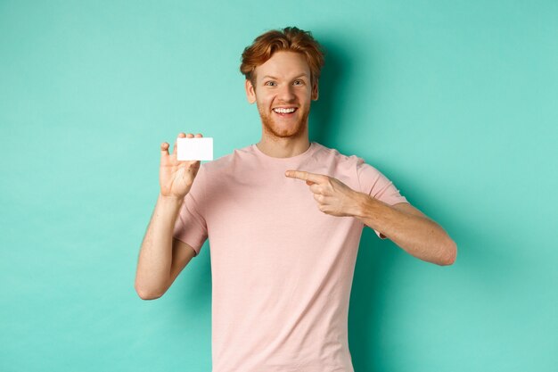 Homme adulte séduisant avec barbe et cheveux roux pointant le doigt sur une carte de crédit en plastique, souriant heureux devant la caméra, debout sur fond turquoise