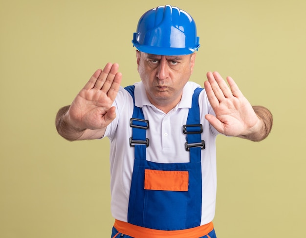 Homme adulte sérieux constructeur en gestes uniformes signe de la main d'arrêt avec deux mains isolé sur mur vert olive