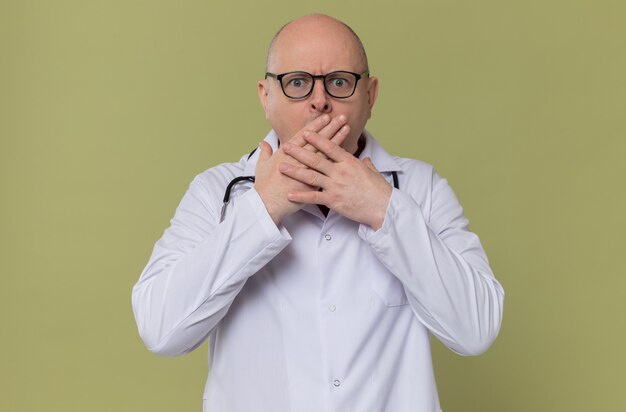 Homme adulte surpris avec des lunettes en uniforme de médecin avec stéthoscope mettant les mains sur sa bouche et regardant