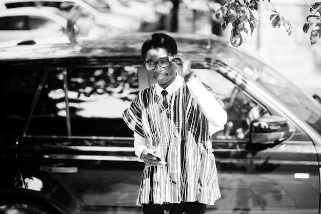 Homme d'affaires africain en vêtements traditionnels et lunettes avec téléphone portable contre voiture noire suv Riches Africains