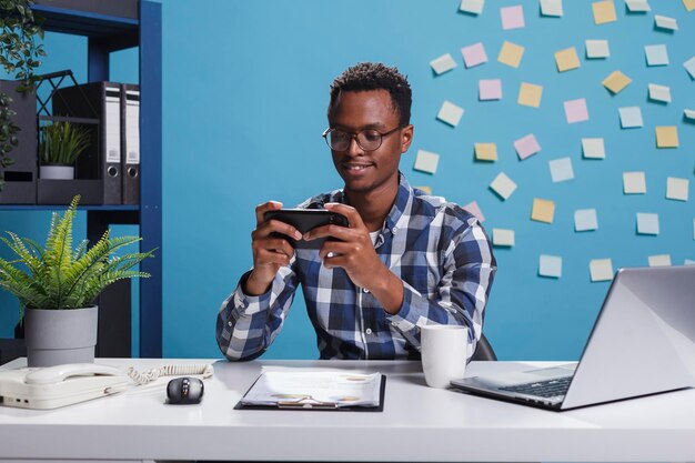 Homme d'affaires afro-américain jouant à des jeux sur un smartphone mobile dans un bureau d'espace de travail moderne. Jeu gagnant du conseiller financier de la société de marketing sur un appareil mobile au travail.