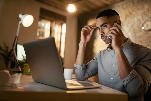 Homme d'affaires afro-américain sérieux passant un appel téléphonique tout en travaillant sur un ordinateur portable la nuit au bureau