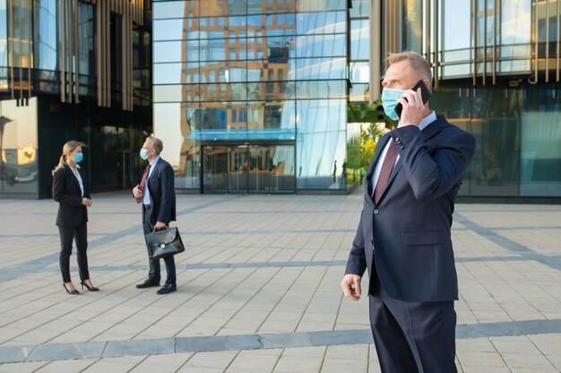 Homme d'affaires d'âge moyen portant masque et costume de bureau, parler sur téléphone mobile à l'extérieur. Les gens d'affaires et la façade en verre du bâtiment de la ville en arrière-plan. Copiez l'espace. Concept d'entreprise et d'épidémie