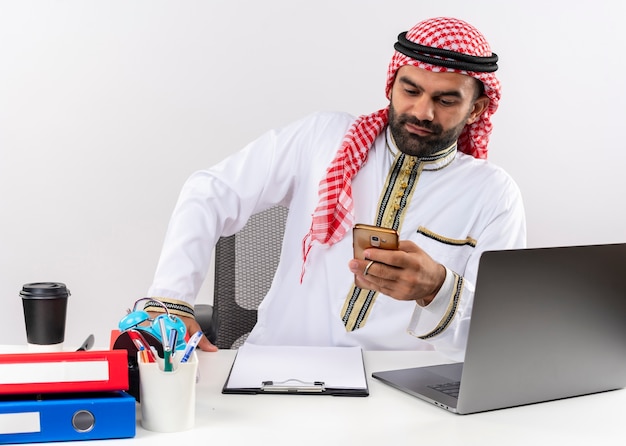 Homme d'affaires arabe en message texto usure traditionnelle sur son smartphone travaillant au bureau lumineux assis à la table