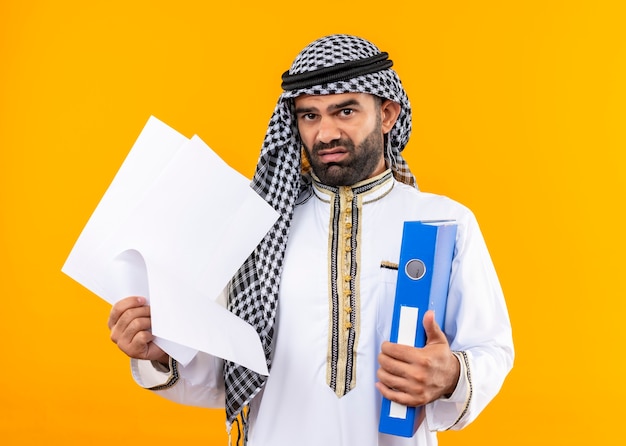 Photo gratuite homme d'affaires arabe en tenue traditionnelle tenant un dossier et des pages blanches mécontent et confus debout sur un mur orange