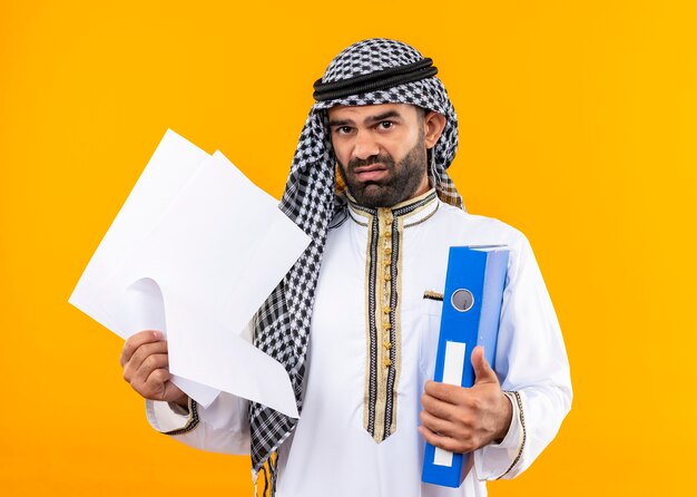 Homme d'affaires arabe en tenue traditionnelle tenant un dossier et des pages blanches mécontent et confus debout sur un mur orange