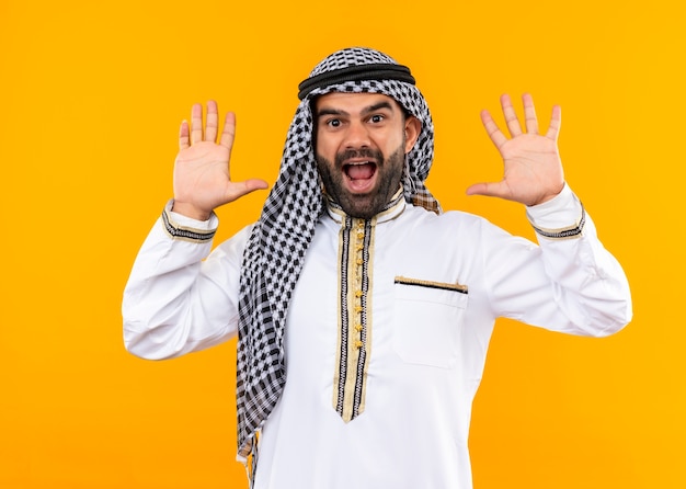 Homme d'affaires arabe en usure traditionnelle en levant les mains dans l'abandon à la surprise debout sur le mur orange