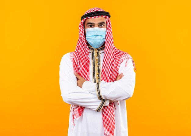 Homme d'affaires arabe en usure traditionnelle et masque de protection du visage avec une expression confiante debout sur un mur orange