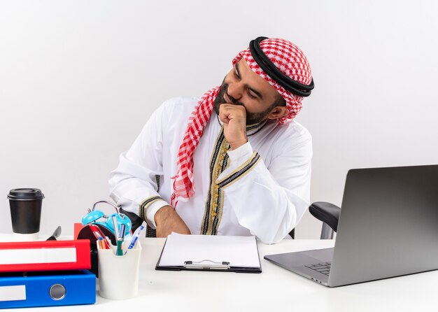 Homme d'affaires arabe en vêtements traditionnels assis à la table avec un ordinateur portable à côté avec une expression pensive perplexe travaillant au bureau