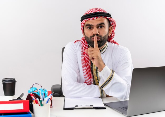 Homme d'affaires arabe en vêtements traditionnels assis à la table avec un ordinateur portable faisant un geste de silence avec le doigt sur les lèvres travaillant au bureau