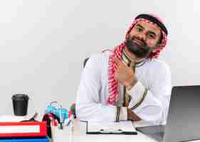 Photo gratuite homme d'affaires arabe en vêtements traditionnels assis à la table avec un ordinateur portable pointant avec l'index vers la gauche à la confiance de travailler au bureau