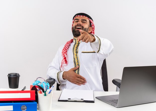 Homme d'affaires arabe en vêtements traditionnels assis à la table avec un ordinateur portable en riant en pointant du doigt pour vous travailler au bureau