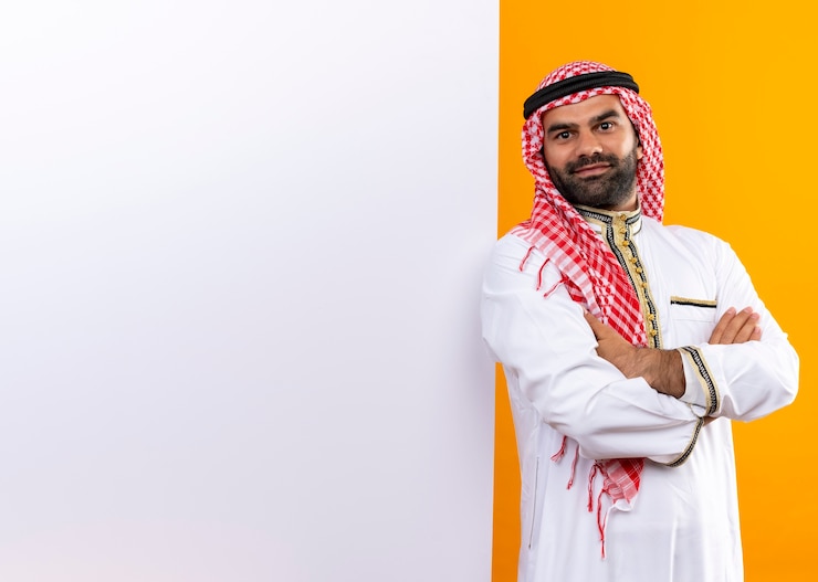 Photo gratuit homme d'affaires arabe en vêtements traditionnels debout près de bilboard vierge avec sourire confiant sur le visage sur le mur orange