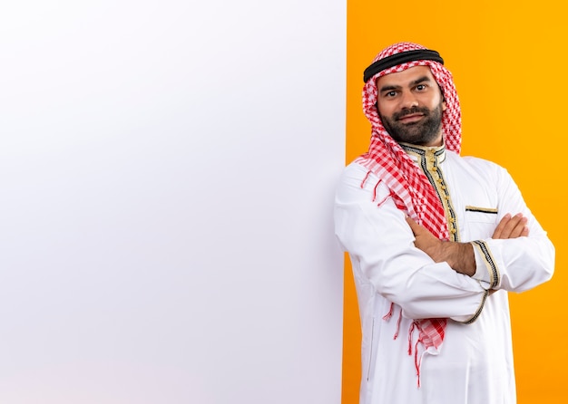 Homme d'affaires arabe en vêtements traditionnels debout près de bilboard vierge avec sourire confiant sur le visage sur le mur orange
