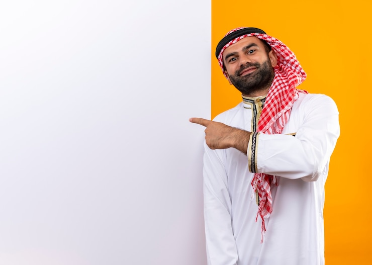Photo gratuit homme d'affaires arabe en vêtements traditionnels debout près de panneau d'affichage vierge pointant avec le doigt dessus avec un sourire confiant sur le visage sur le mur orange